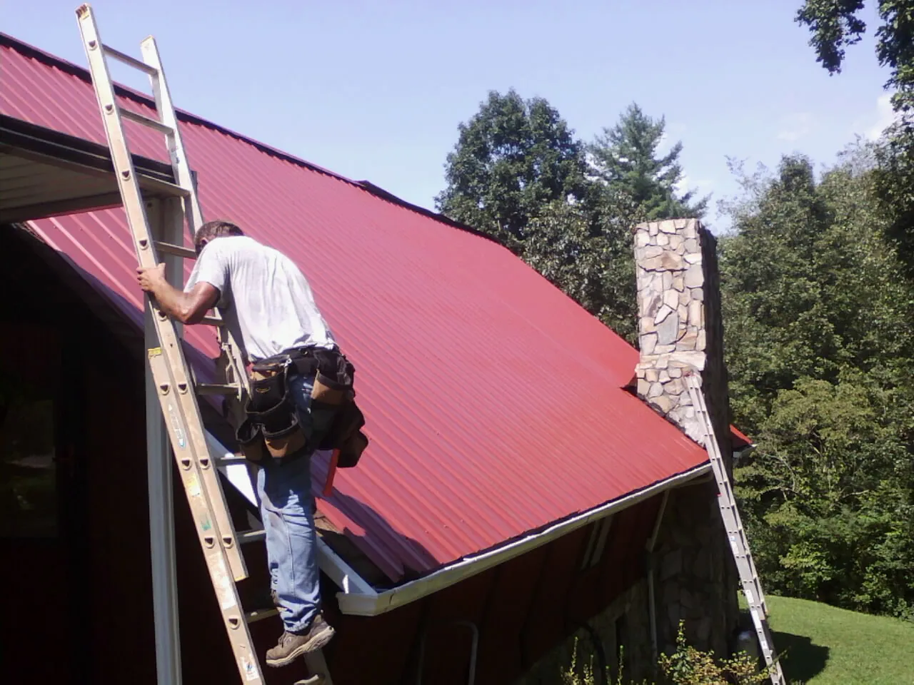 Roofer on a ladder beside a red metal roof and stone chimney — Soffit & Fascia Repair by Roofing Brothers in Menlo
